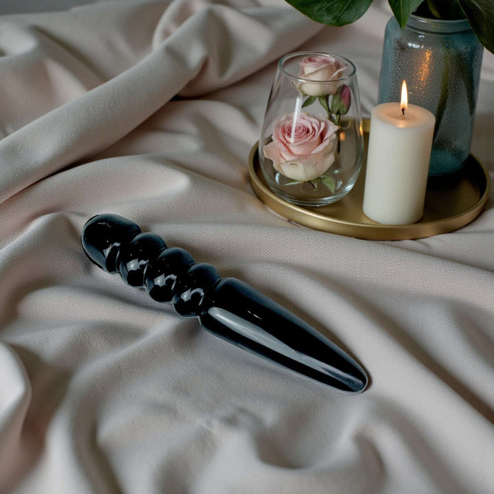 Black Obsidian crystal pleasure wand laying on a white sheet with botanicals 
