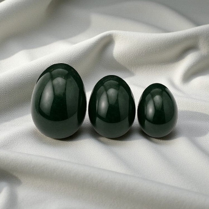 Three jade yoni eggs of varying sizes placed on a soft white fabric, showcasing their smooth, polished surface.